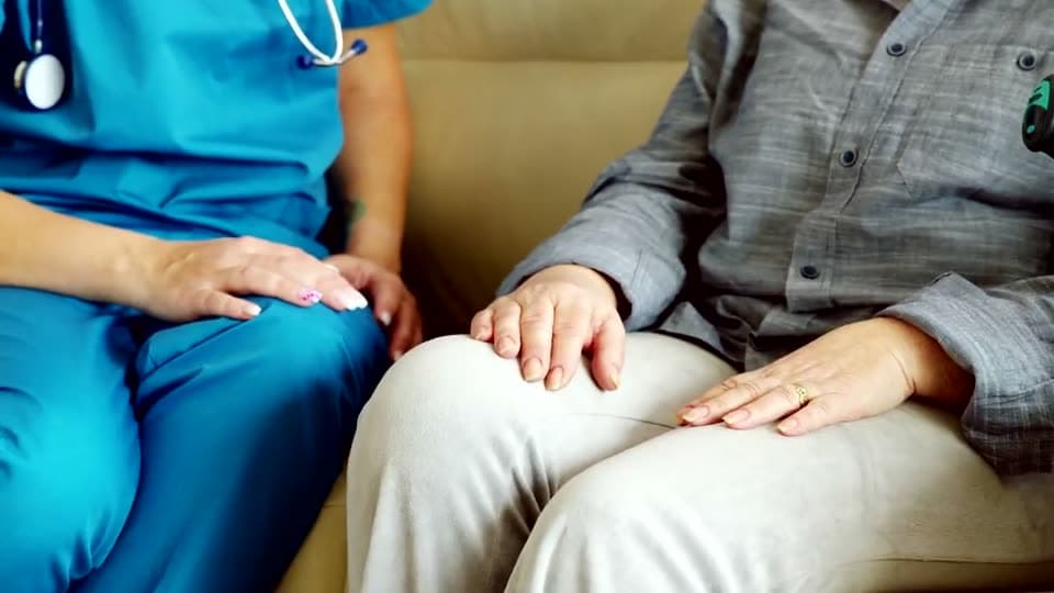 A nurse rests both hands over the hands of an older person seated beside her on a sofa.