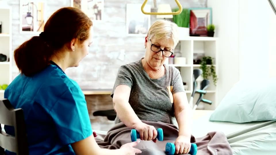A nurse steadies the arm of an older woman as she lifts a small dumbbell during an exercise session at home.