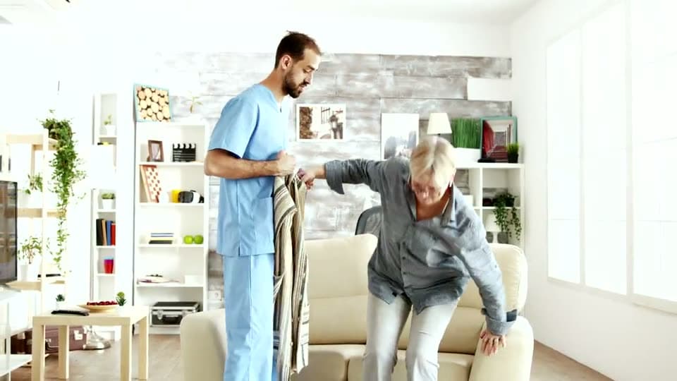 A nurse helps an older woman into her cardigan as she steadies herself on her feet beside an armchair.