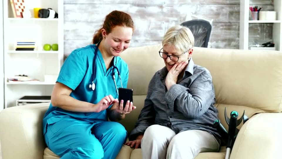 A home health nurse and an older woman sit together on a sofa, sharing something on a phone and smiling.