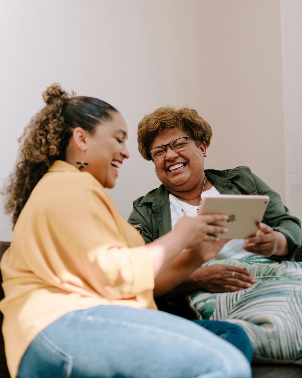 Two women sit together on a sofa in a bright living room, laughing over a tablet the older woman is holding.