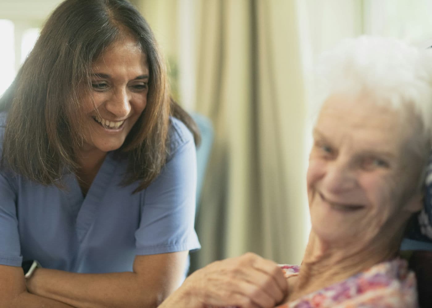 A nurse in blue scrubs leans in and smiles with an older woman by a sunlit window at home, their hands together.